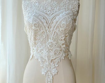 May include: An ivory-colored lace applique featuring intricate floral and scroll embroidery with delicate beadwork. The design is symmetrical, tapering to a point at the bottom, and is displayed on a mannequin to show the detailed needlework.