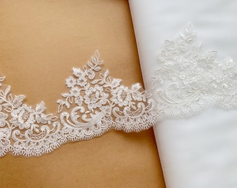 May include: A white lace trim featuring an intricate floral and scroll pattern with scalloped edges. The lace is displayed on a light brown surface and partially layered over a sheer white fabric, highlighting the detailed embroidery and stitching.