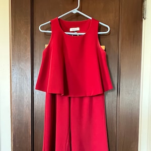 May include: A red sleeveless jumpsuit with a ruffled top and wide-leg pants. The jumpsuit is hanging on a white hanger against a wooden door.