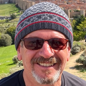 May include: A knit beanie featuring a gray and white geometric pattern with a solid dark gray ribbed brim and a thin horizontal red stripe. The hat is constructed from yarn and sits snugly on the head, providing full coverage.