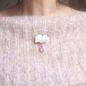 May include: A gold-tone chain necklace with a pendant depicting an open book and a teardrop charm. The book pendant is light-coloured with printed text, and the charm has a pink, green, and yellow pattern.