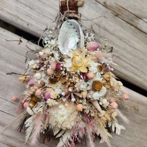 May include: A handcrafted bouquet featuring dried flowers in shades of cream, pink, and brown, accented with a pearlescent abalone shell. The arrangement is tied with natural twine and includes delicate sprigs and seed pods, creating a rustic, natural aesthetic.