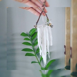 May include: A small white fabric dream catcher hanging from a twig triangle with dried flowers. The dream catcher is made of white fabric strips and is hanging from a twig triangle. The dream catcher is hanging from a twig triangle with dried flowers.