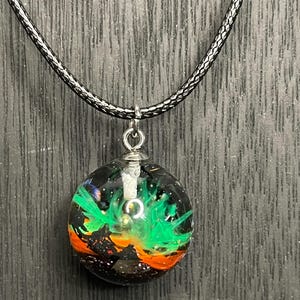 May include: A round pendant necklace with a black cord. The pendant is a clear orb with a colorful design inside, featuring green, orange, and black elements. The design resembles a nebula or abstract landscape.