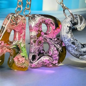 May include: Three resin keychains shaped as the letters A, B, and C. The letter A features swirls of pink, green, and amber. The letter B contains pink and black marbled patterns. The letter C displays a clear, black, and white marbled effect.