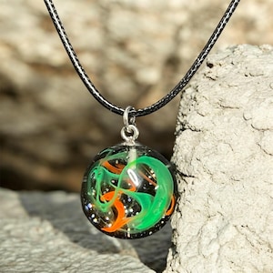 May include: A round pendant necklace with a black cord. The pendant is a clear orb filled with swirling orange and green shapes, and tiny glitter particles. The pendant is attached to a silver-colored bail.