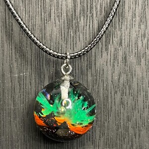 Puede incluir: Collar con colgante redondo y cordón negro. El colgante transparente contiene un diseño colorido con elementos verdes, naranjas y negros, que evoca una nebulosa o arte abstracto.