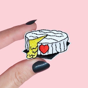 Peut inclure: Patch brodé blanc à repasser représentant un rond de brie avec une part enlevée et un cœur rouge sur le côté. Le fromage coule avec du fromage jaune.