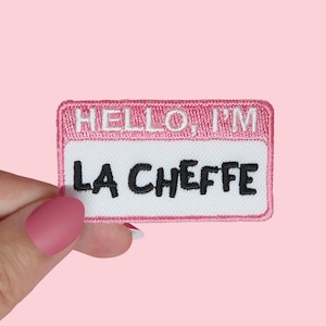 Puede incluir: Parche de tela rosa con costuras blancas y texto negro que dice "HELLO, I'M LA CHEFFE" con la bandera francesa y "MADE IN FRANCE" debajo.
