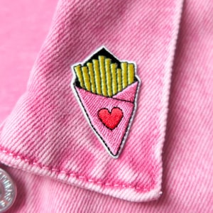 Peut inclure: Une veste en jean rose avec un petit patch brodé représentant un contenant de frites en forme de cœur. Le contenant est rose avec un cœur rouge et des frites jaunes.