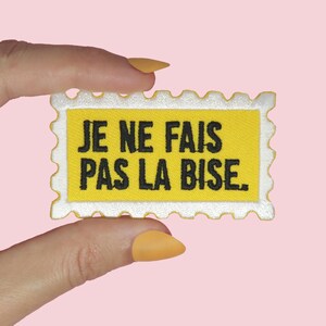 Peut inclure: Un écusson brodé rectangulaire en forme de timbre-poste avec un centre jaune et une bordure blanche. Le texte JE NE FAIS PAS LA BISE est brodé en lettres capitales noires au centre de l'écusson sur le fond jaune.