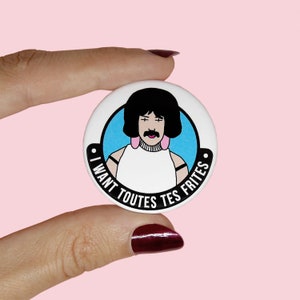 Può includere: Un bottone bianco con un cerchio blu e un'immagine di cartone animato di Freddie Mercury con il testo "I WANT TOUTES TES FRITES" attorno al cerchio.