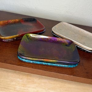 May include: Three rectangular glass dishes with iridescent finishes. The dishes are stacked on top of each other. The top dish is a deep purple with a turquoise rim. The middle dish is a deep red with a gold rim. The bottom dish is a light beige with a black rim.