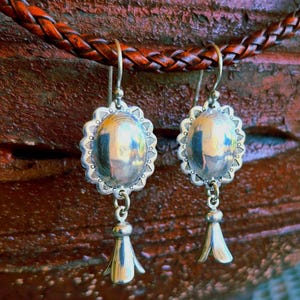 May include: A pair of silver earrings with an oval, scalloped design. Each earring has a small, dangling bell-shaped charm. The earrings are displayed against a brown, braided leather background.