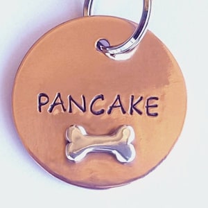 May include: Copper dog tag with the name "Pancake" engraved on it and a silver bone charm attached.