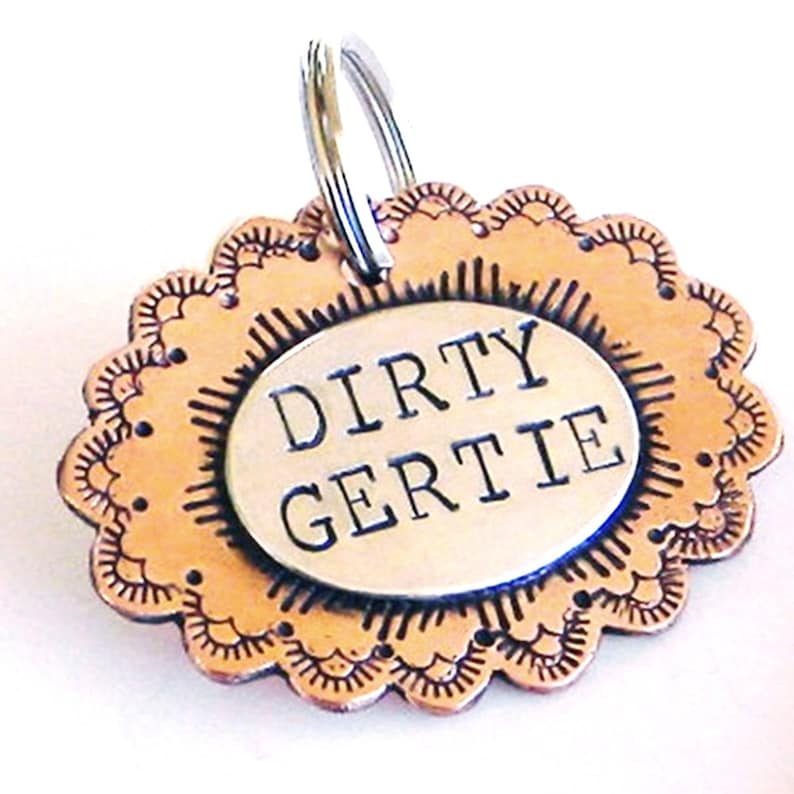 May include: A copper-colored dog tag with a scalloped edge and a silver oval charm that reads "DIRTY GERTIE".