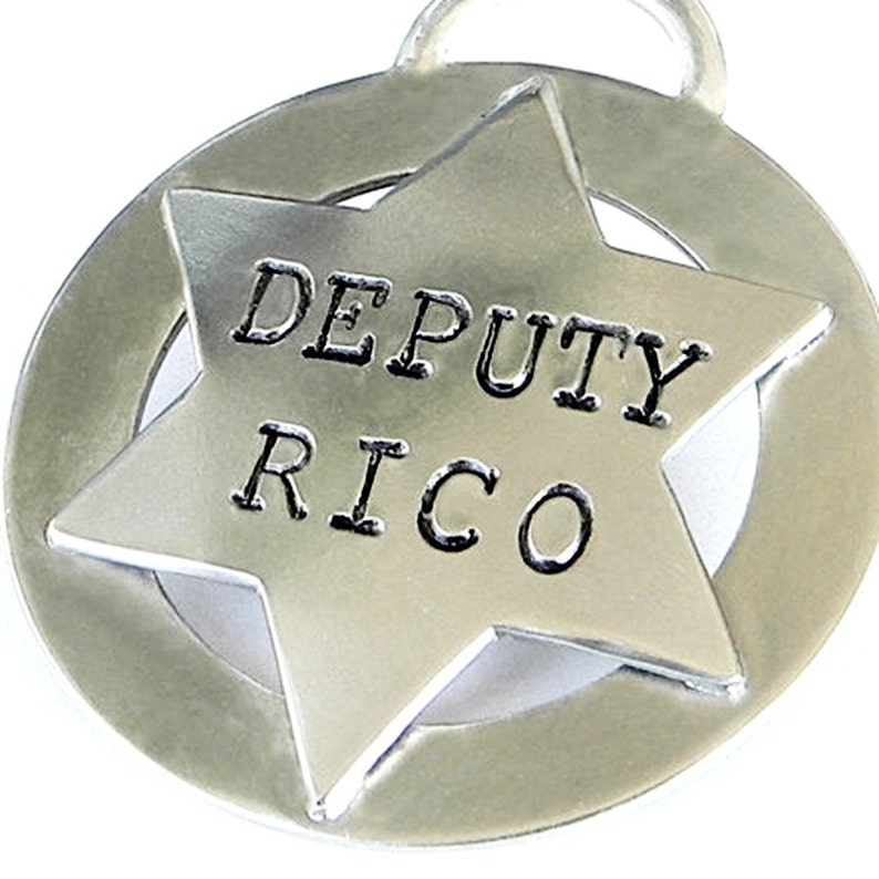 May include: A silver-colored pet tag in the shape of a sheriff's star with the words "DEPUTY RICO" engraved on it. The star is set within a circular frame, and a small loop is at the top for attachment to a collar.