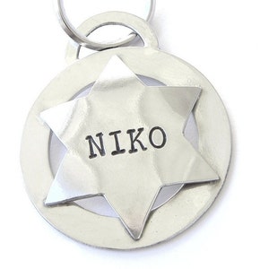 May include: A silver pet tag with the name "NIKO" stamped on a six-pointed star. The star is set within a circular frame, and a metal ring is attached for easy attachment to a collar. The tag is made of shiny, polished metal.