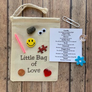 May include: A beige canvas bag with a drawstring closure, labeled "Little Bag of Love." The bag is adorned with a variety of small items, including a candle, a smiley face button, a puzzle piece, and a red heart.