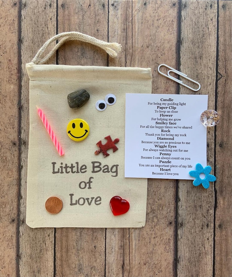 May include: A beige canvas bag with a drawstring closure, labeled "Little Bag of Love." The bag is adorned with a variety of small items, including a candle, a smiley face button, a puzzle piece, and a red heart.