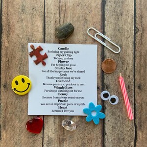 May include: A collection of small, symbolic items arranged on a wooden surface. Includes a red heart, a smiley face magnet, a penny, a paperclip, a candle, a puzzle piece, a flower, a rock, a diamond, and googly eyes. A card lists each item with a short message.