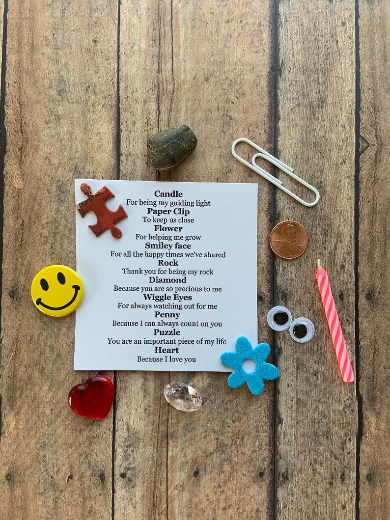 May include: A collection of small, symbolic items arranged on a wooden surface. Includes a red heart, a smiley face magnet, a penny, a paperclip, a candle, a puzzle piece, a flower, a rock, a diamond, and googly eyes. A card lists each item with a short message.
