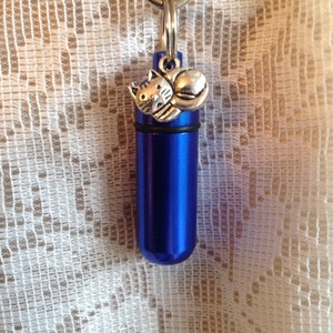 May include: A blue metal keychain with a small silver cat charm. The keychain is designed to hold a small amount of ashes or other mementos.