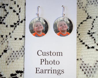 May include: A pair of custom photo earrings. Each earring features a circular design with a photo of a smiling child. The earrings hang from silver-colored hooks. The card reads "Custom Photo Earrings".
