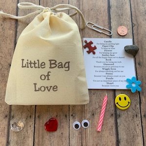 May include: A beige canvas bag with the text "Little Bag of Love" sits on a wooden surface, surrounded by small trinkets. These include a paperclip, a penny, a red heart, a candle, a smiley face, and a small card with a poem.