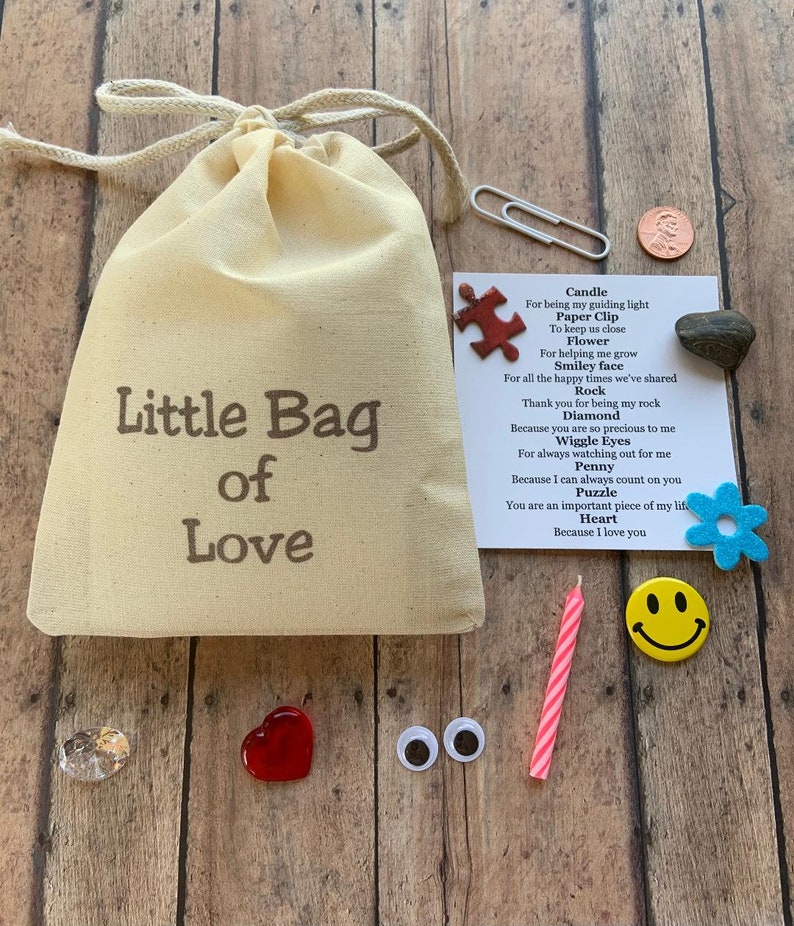May include: A beige canvas bag with the text "Little Bag of Love" sits on a wooden surface, surrounded by small trinkets. These include a paperclip, a penny, a red heart, a candle, a smiley face, and a small card with a poem.