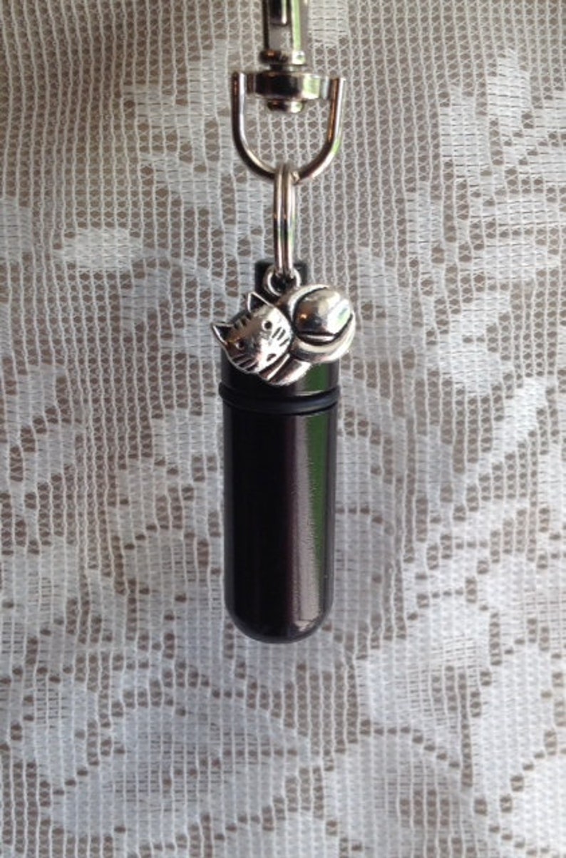 May include: A black metal keychain with a small silver charm of a cat holding a ball. The keychain has a loop for attaching to a bag or belt.