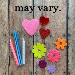 May include: Assortment of decorative items including glittery hearts, colorful flower shapes, and striped candles. The text "Some items may vary" is displayed at the top of the image. The items are arranged on a wooden background.