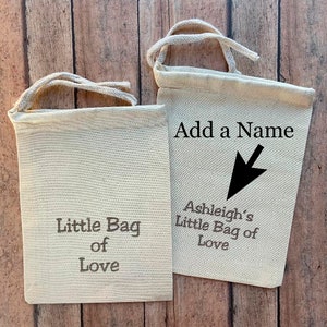May include: Two small, off-white canvas bags with drawstring closures. One bag reads "Little Bag of Love" in gray text. The other bag has the text "Add a Name" with an arrow pointing to "Ashleigh's Little Bag of Love".