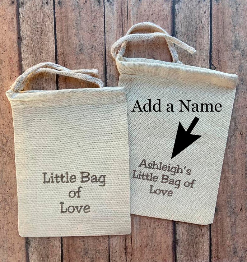 May include: Two small, off-white canvas bags with drawstring closures. One bag reads "Little Bag of Love" in gray text. The other bag has the text "Add a Name" with an arrow pointing to "Ashleigh's Little Bag of Love".