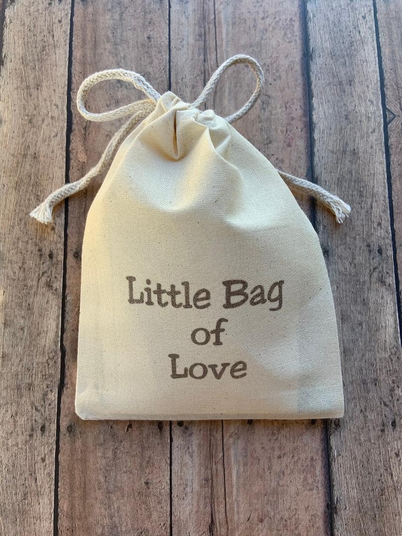 May include: A small, cream-colored drawstring bag with the text "Little Bag of Love" printed in brown. The bag is made of a soft fabric and has a natural, rustic appearance. The background is a wooden surface.
