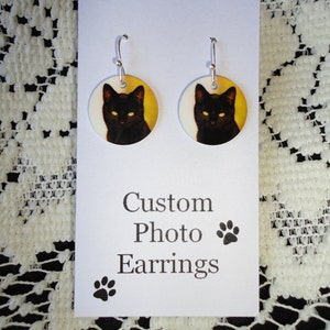 May include: A pair of custom photo earrings featuring a black cat with yellow eyes on a yellow background. The earrings are round and have silver hooks. The card reads "Custom Photo Earrings" with a paw print.