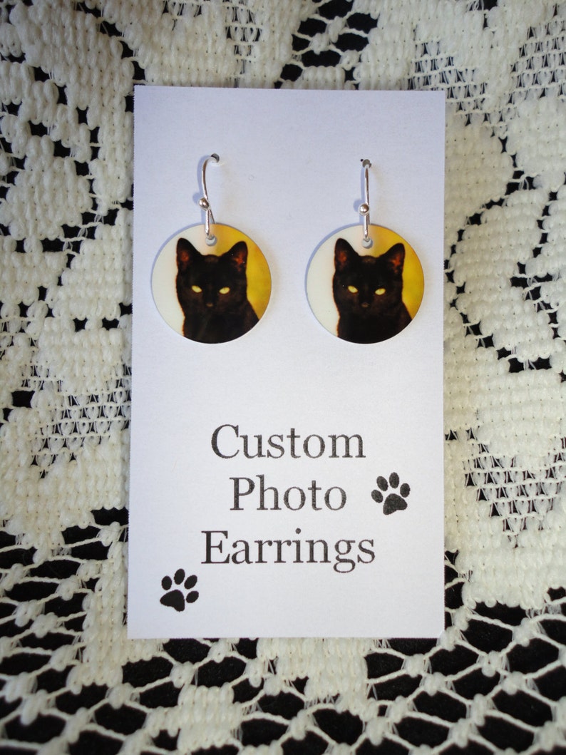 May include: A pair of custom photo earrings featuring a black cat with yellow eyes on a yellow background. The earrings are round and have silver hooks. The card reads "Custom Photo Earrings" with a paw print.