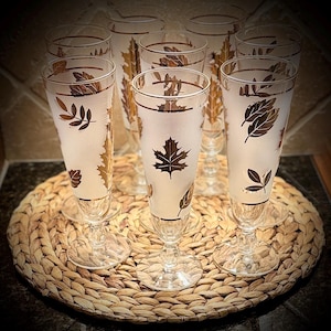 May include: Set of vintage champagne flutes with frosted glass and gold leaf designs. The glasses are arranged on a woven placemat. Each glass features different leaf patterns, adding a touch of elegance to any occasion.