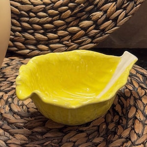 May include: A bright yellow ceramic bowl shaped like a cabbage leaf. The bowl features a glossy finish with realistic, textured ridges and veins that mimic the appearance of a vegetable leaf, including a prominent stem detail on the side rim.