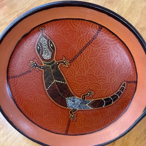 May include: A round, orange decorative plate with a black rim and a lizard design. The lizard is black, brown, and white, with a patterned body and a dotted pattern around it. The background has a repeating wave pattern.