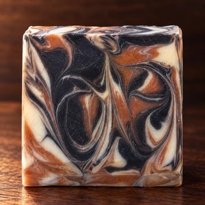May include: A rectangular bar of soap featuring a marbled swirl pattern in shades of black, copper, and cream. The surface of the soap is smooth and flat, displaying a complex, flowing design created by the blending of these three distinct colours.