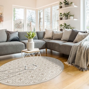 May include: A round, beige and white patterned area rug with a geometric design. The rug is placed in a living room with a gray sectional sofa and a white coffee table.