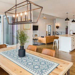 May include: A light wood dining table with a blue and white patterned table runner. The table is set with four chairs and a vase of greenery. The table is in a modern dining room with a kitchen in the background.