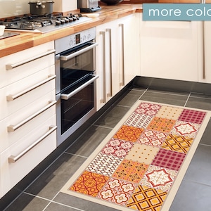 Puede incluir: Una alfombra de cocina con un diseño de patchwork con azulejos rojos, naranjas, amarillos y blancos. La alfombra está delante de un armario de cocina blanco con electrodomésticos de acero inoxidable.