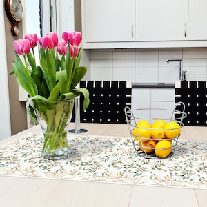 May include: A white table runner with a green and gold leaf pattern. A bouquet of pink tulips in a clear glass vase and a metal basket filled with yellow lemons are on the table runner.