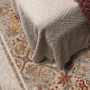 May include: A close-up of a beige patterned rug with a white crocheted blanket draped over it. The blanket has a diamond pattern and is folded over at the edges.
