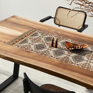 May include: A large, rectangular wooden dining table with a decorative runner featuring a geometric pattern in beige, blue, and brown. Two wooden candle holders and a wooden bowl with nuts are on the table. Black chairs with woven backs are visible.