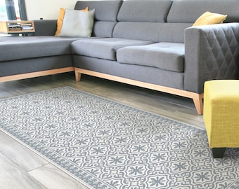 Puede incluir: Una alfombra de área gris y blanca con un diseño geométrico. La alfombra se coloca frente a un sofá seccional gris con una almohada amarilla.
