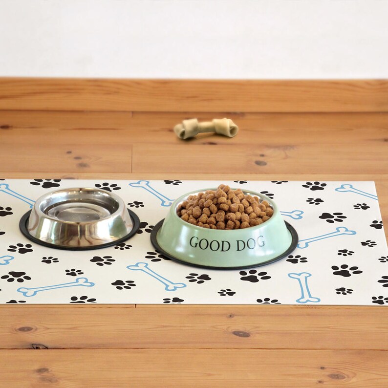 Op de afbeelding: Een witte hondenvoermat met zwarte poot afdrukken en blauwe botmotieven. Twee roestvrijstalen kommen staan op de mat, een gevuld met hondenvoer en de andere leeg. De groene kom heeft de tekst "GOOD DOG" erop gedrukt.