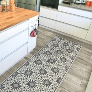 May include: A white and gray patterned kitchen runner rug with a geometric floral design. The rug is placed in a kitchen with white cabinets and a wood floor.